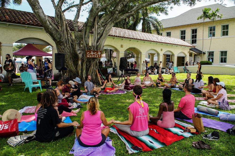 A large circle of people sitting on mats and blankets on the lawn under the banyan tree at Old School Square, many wearing pink, during a session at Coco Market Goes Pink.