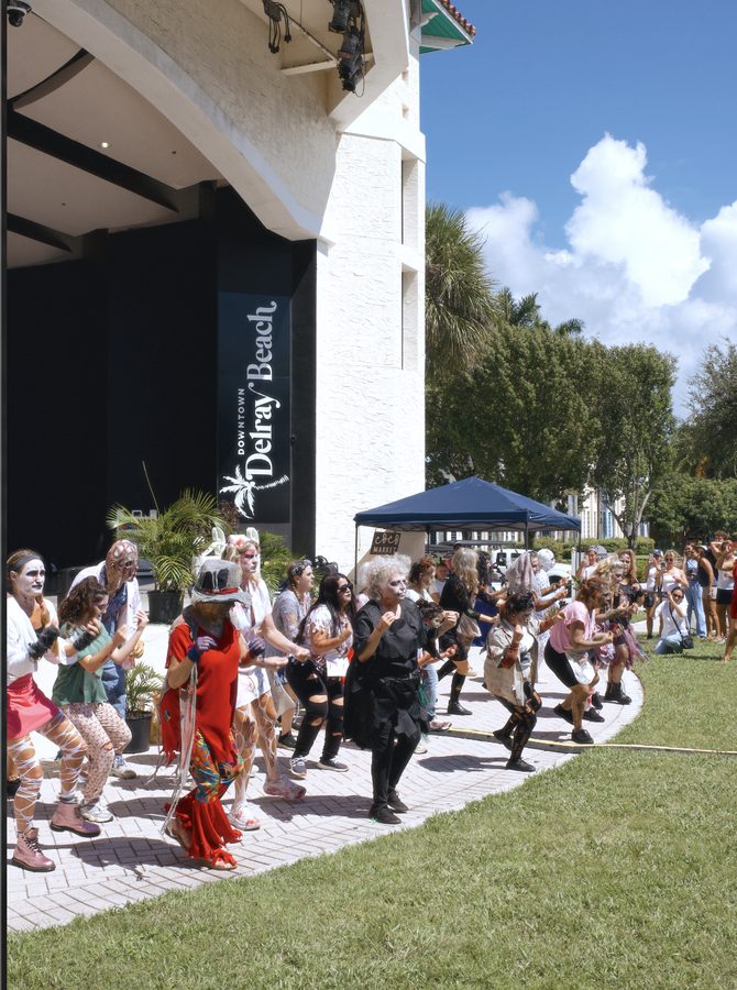 Dancers in zombie costume and make-up moving together in front of the Delray Beach sign.