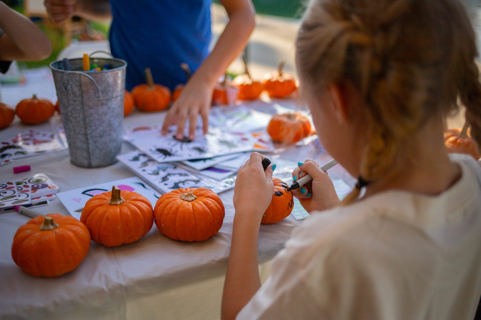A child decorating a mini pumpkin with a black marker at a craft table lined with pumpkins, sticker sheets and a tin of pens.